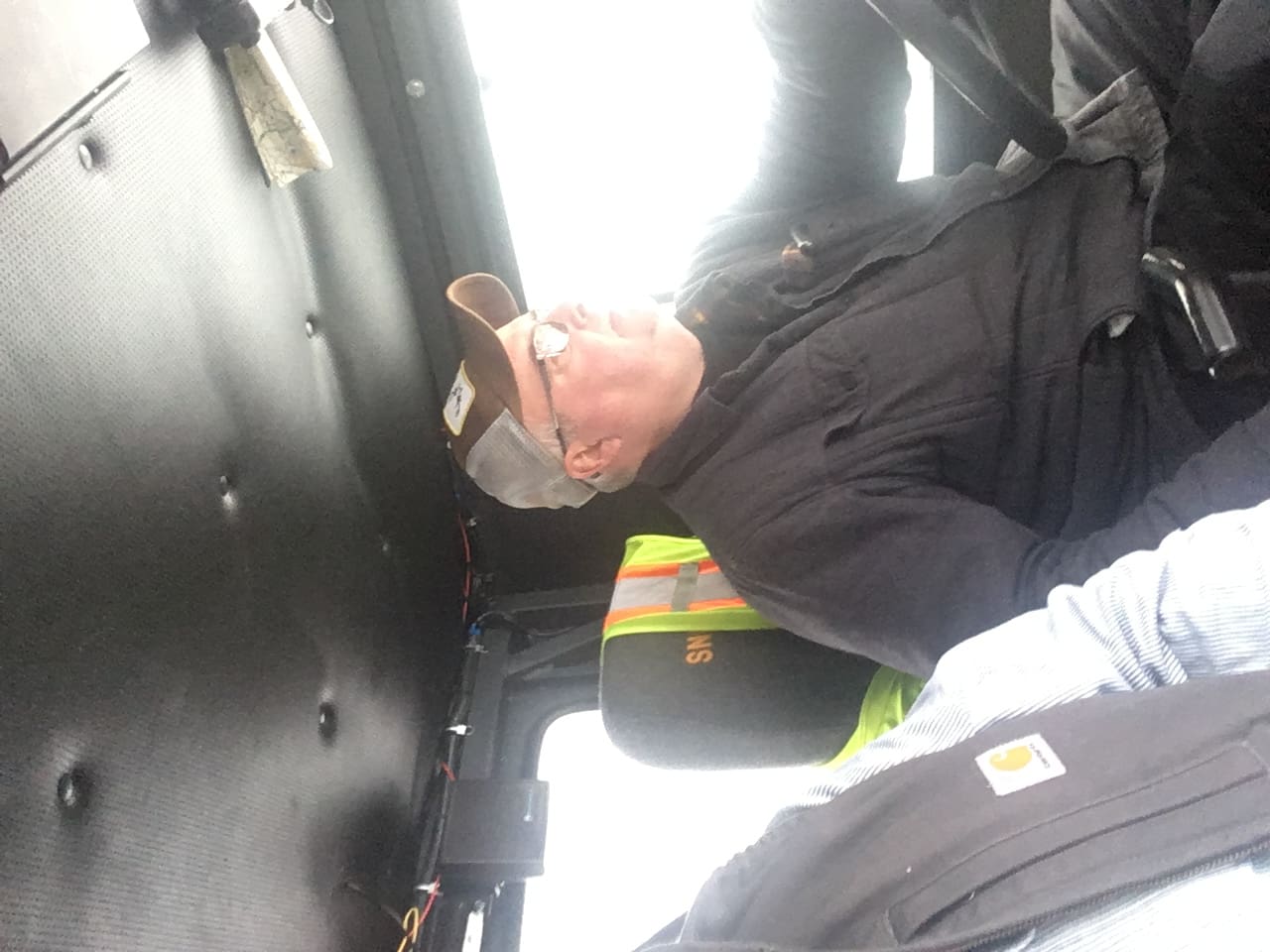 An upside-down image of a man wearing a hat and safety vest, seated and seemingly asleep inside a vehicle.