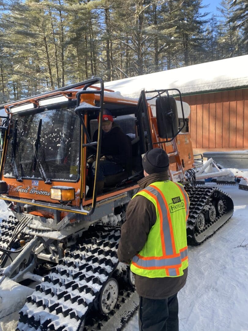 A person wearing a visibility vest talks to another person inside an orange snow groomer in a snowy forest setting.