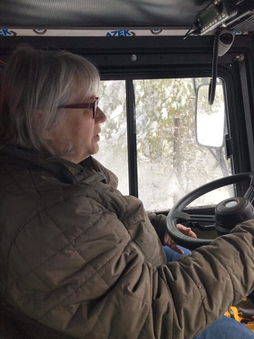A woman wearing glasses and a green jacket driving a vehicle in snowy conditions, viewed from the side.
