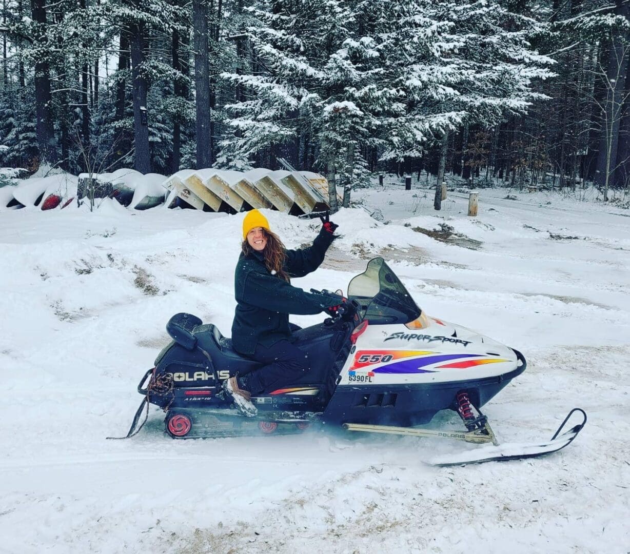 A woman wearing a yellow hat sits on a snowmobile, waving, in a snowy forest setting.