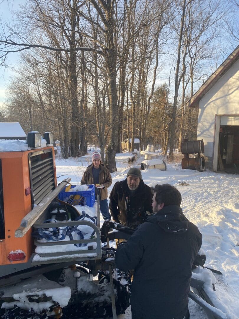 Three people working on a snow-covered vehicle next to a building, surrounded by snowy trees and equipment.