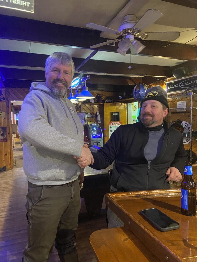 Two men smiling and shaking hands at a bar with a beer and a phone on the table in front of them.