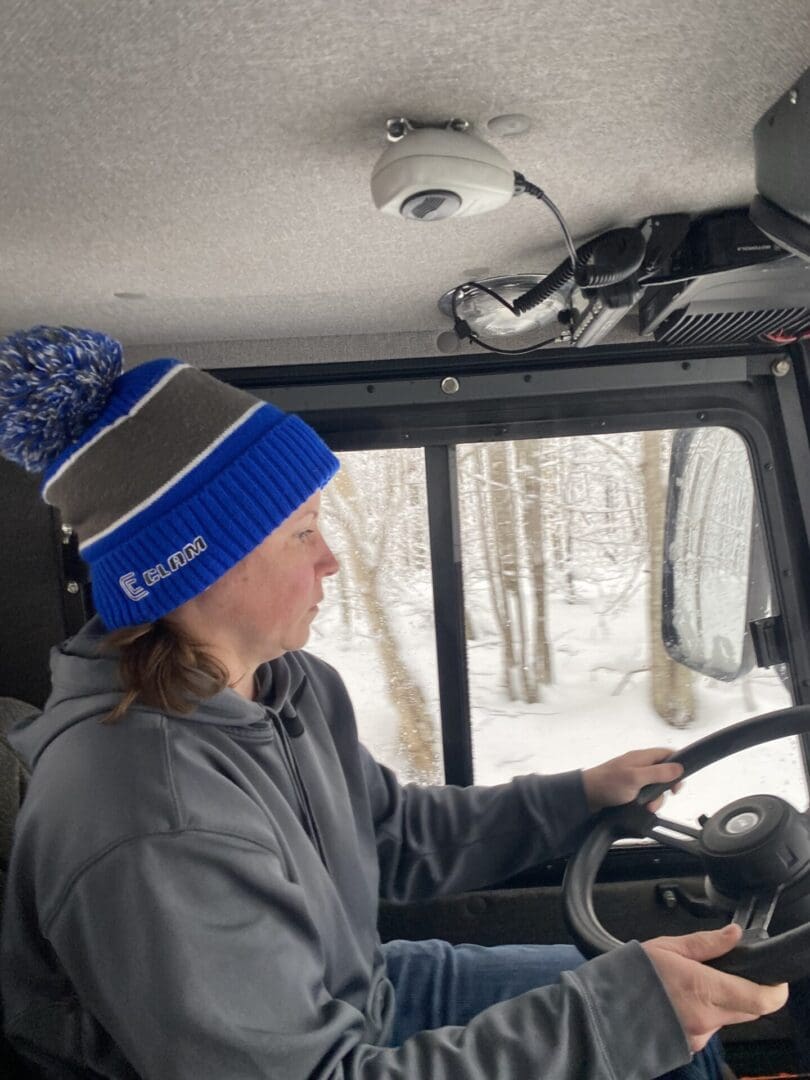 A woman driving a snow-covered vehicle, wearing a blue beanie and gray hoodie, focused on the road with snowy trees visible outside.