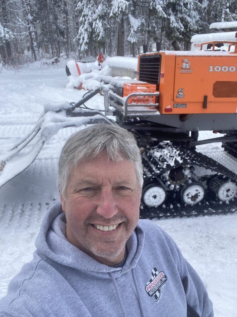 Man smiling for a selfie in a snowy setting with a large orange snow grooming machine in the background.