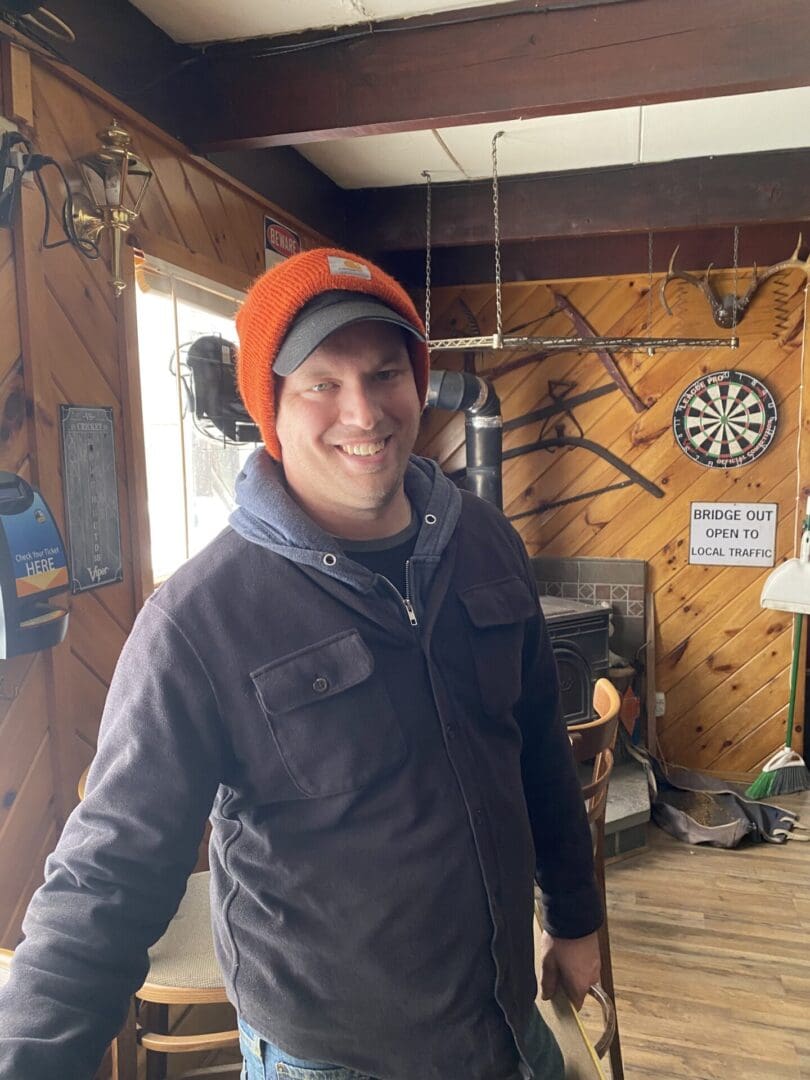 Man in a blue jacket and orange beanie smiling in a rustic room with wood paneling and a dartboard in the background.