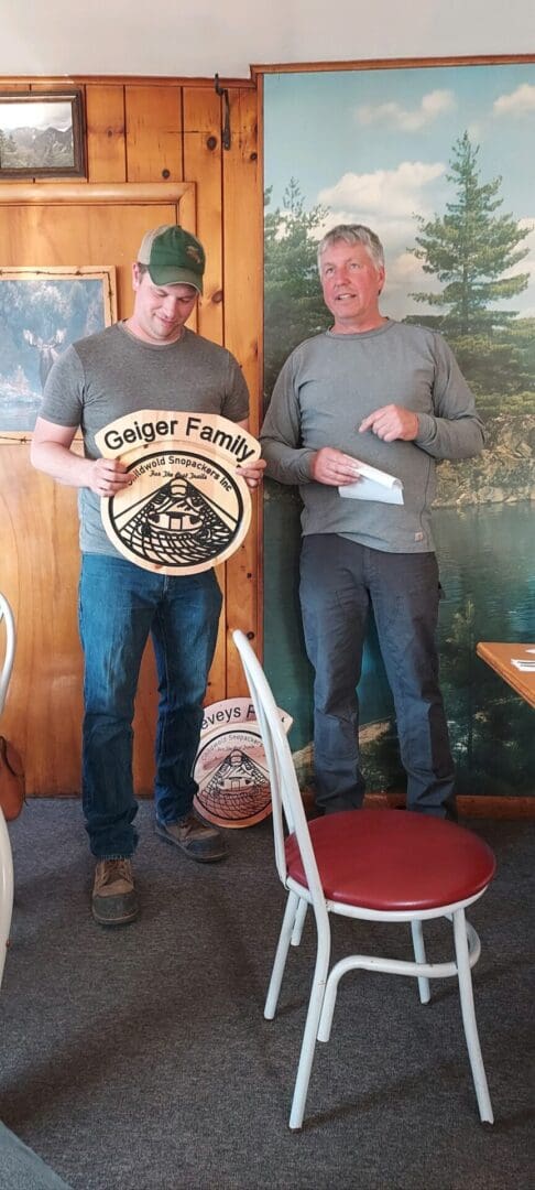 Two men standing indoors, one holding a circular wooden sign with 