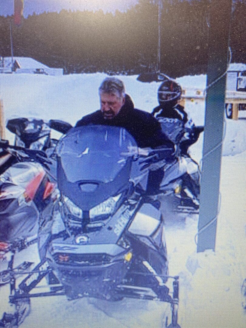 Two people on snowmobiles parked in a snowy area, one in the foreground looking towards the camera, and another slightly behind wearing a helmet.