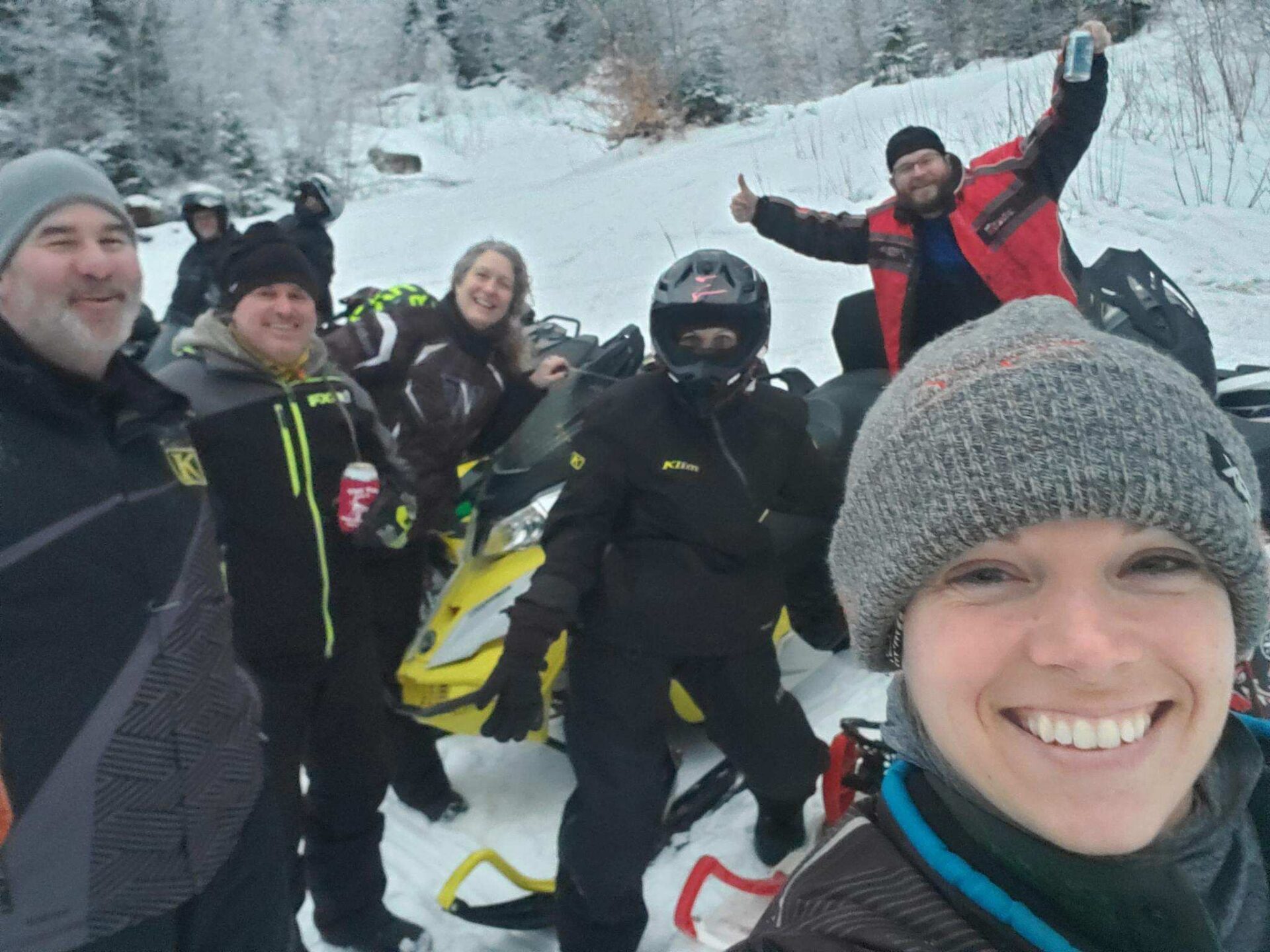 A group of six people posing with snowmobiles in a snowy landscape, smiling and raising their hands in celebration.