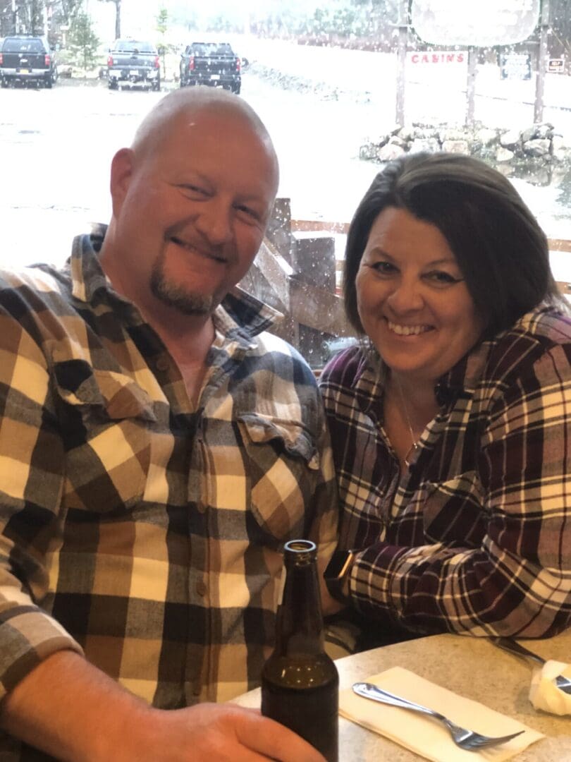 A man and a woman in plaid shirts smiling at a table with a bottle in front, in a cabin-style restaurant with visible snow outside.
