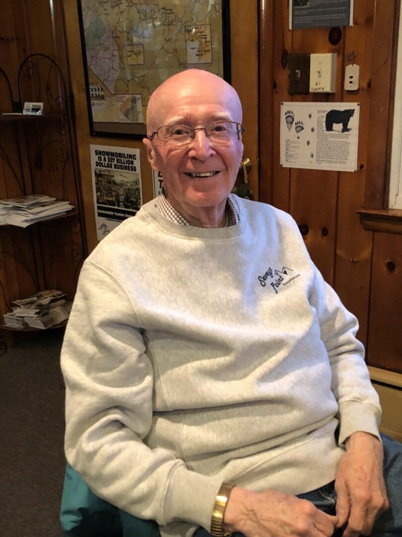 Elderly man smiling in a wheelchair, wearing glasses and a light gray sweatshirt, inside a room with maps and posters on the wall.