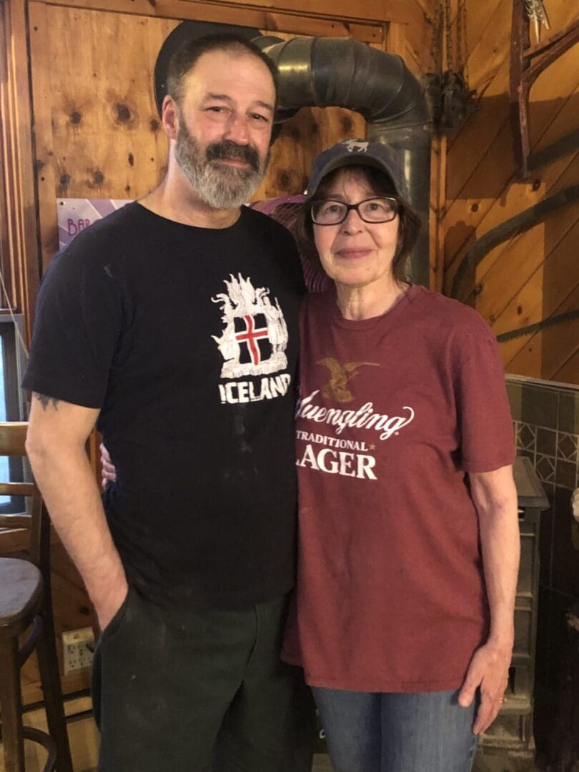 A middle-aged man and woman smiling inside a rustic wooden room, wearing t-shirts and standing close together.