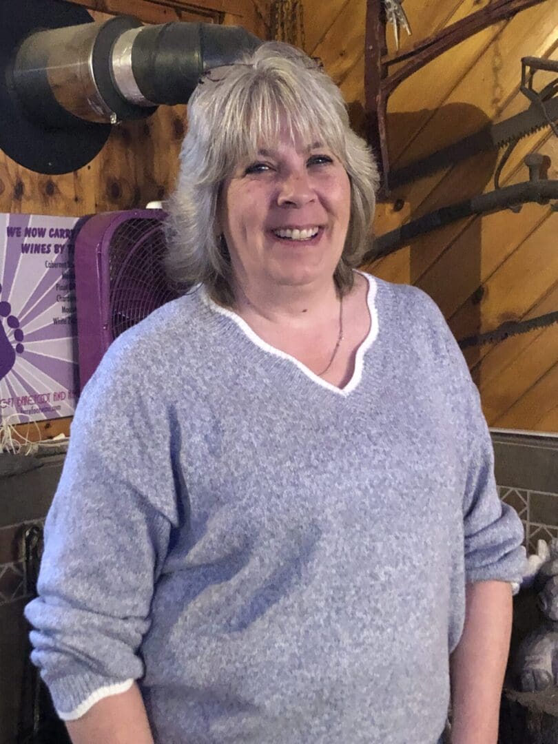 A middle-aged woman smiling, wearing a blue sweater, standing in a rustic room with wood paneling and a purple fan in the background.