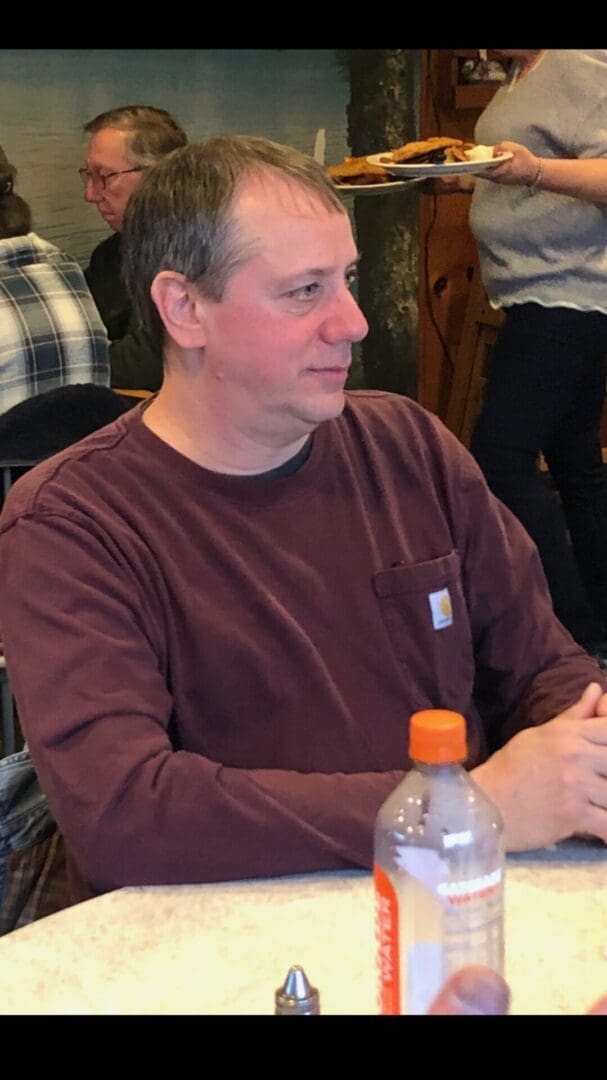 Man in a burgundy shirt sitting at a table with a bottle in front, in a crowded room with focus on him.