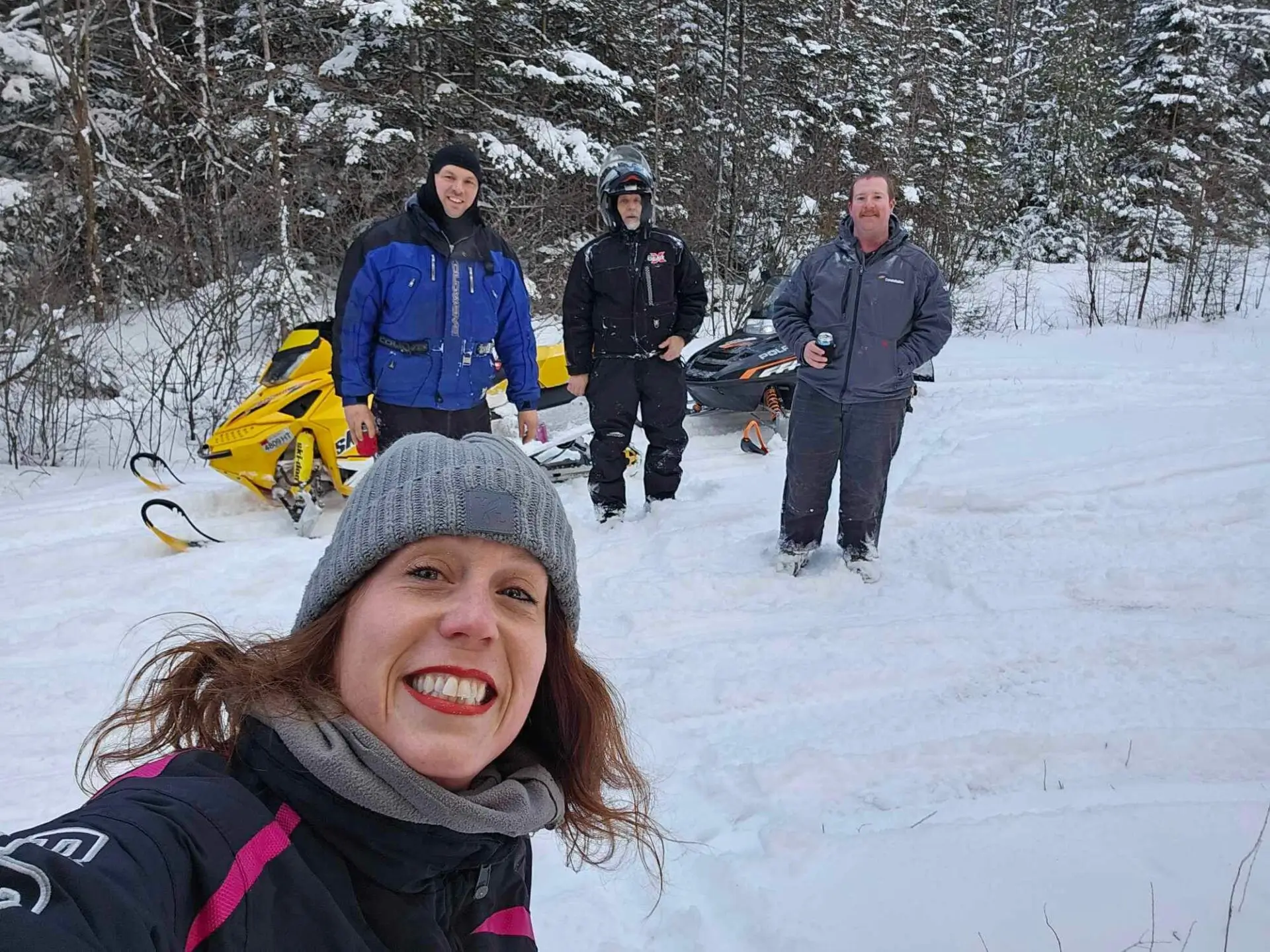 A woman in the foreground takes a selfie with three men standing behind her in a snowy landscape, with snowmobiles visible.