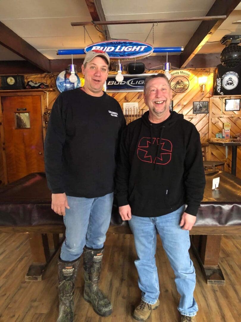 Two men smiling inside a rustic bar, standing under a bud light sign, one in a black hoodie, the other in a gray sweatshirt.