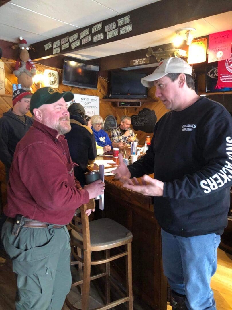 Two men talking at a bar, one holding a drink, surrounded by other patrons and sports memorabilia.