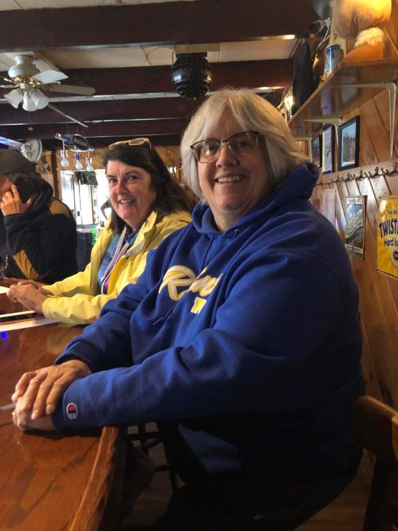 Two women smiling at a bar, one in a blue hoodie and the other in a yellow jacket, surrounded by wooden decor and hanging lights.