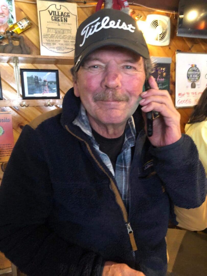 An older man with a mustache, wearing a titleist cap and a blue fleece jacket, talks on a mobile phone in a busy indoor setting.