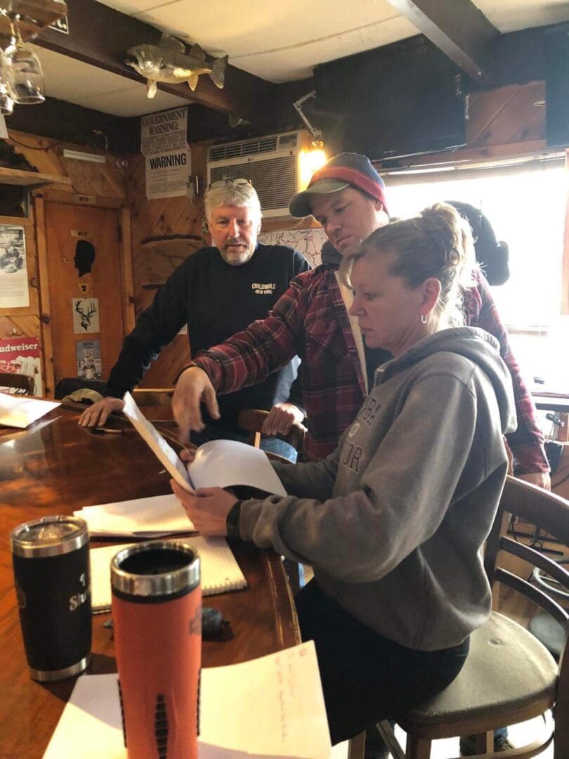 Three people discussing a document in a pub with rustic decor, including two men and a woman, surrounded by various signs and beverage containers.